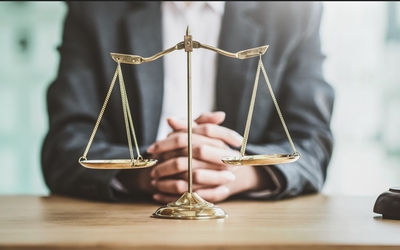 Photo d'un avocat assis à son bureau, joignant ses mains entre elles. Devant lui se trouve une balance (symbole de la justice) et un marteau