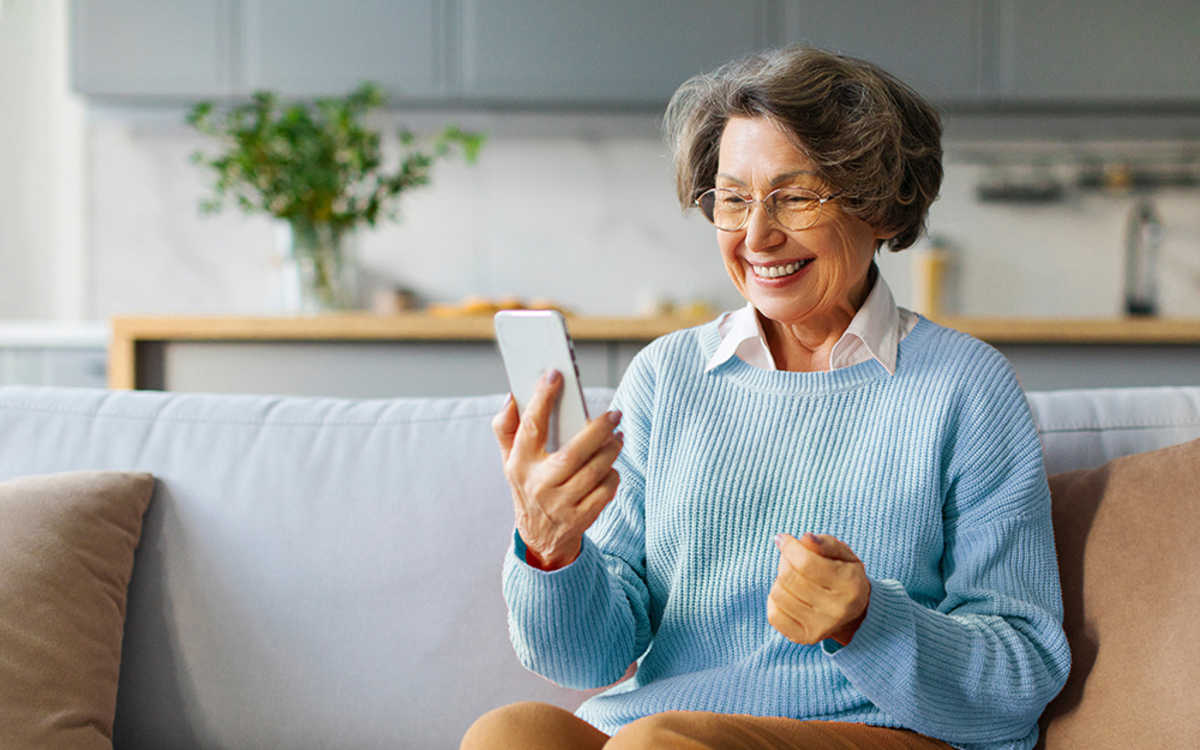 Une personne âgée est assise sur son canapé et sourit en regardant son smartphone.