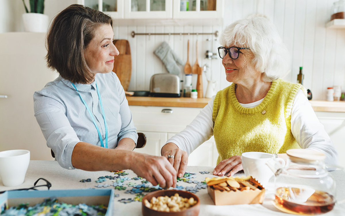 Photographie montrant une jeune femme qui aident une personne âgée à faire un puzzle. Elles sourient toutes les deux. Elles ont assises à une table à manger.
