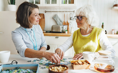 Photographie montrant une jeune femme qui aident une personne âgée à faire un puzzle. Elles sourient toutes les deux. Elles ont assises à une table à manger.