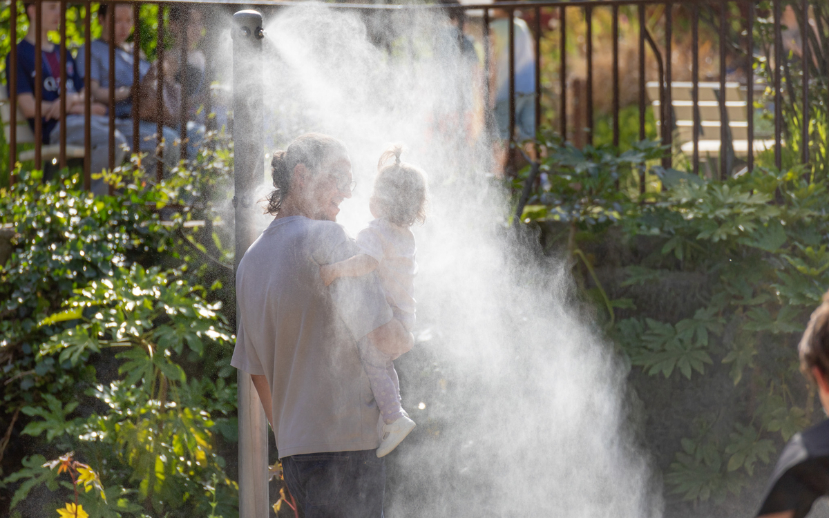 Un adulte avec un enfant dans les bras se rafraîchissent sous un brumisateur dans un parc