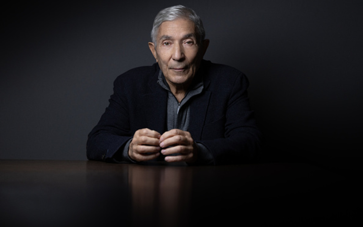 Photo de boualem Saensal un gomme aux cheveux courts gris regardant la caméra de face vetu d'un pull gris et une veste noire, sur un fond noir les mains posées sur la table grise