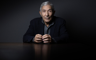 Photo de boualem Saensal un gomme aux cheveux courts gris regardant la caméra de face vetu d'un pull gris et une veste noire, sur un fond noir les mains posées sur la table grise