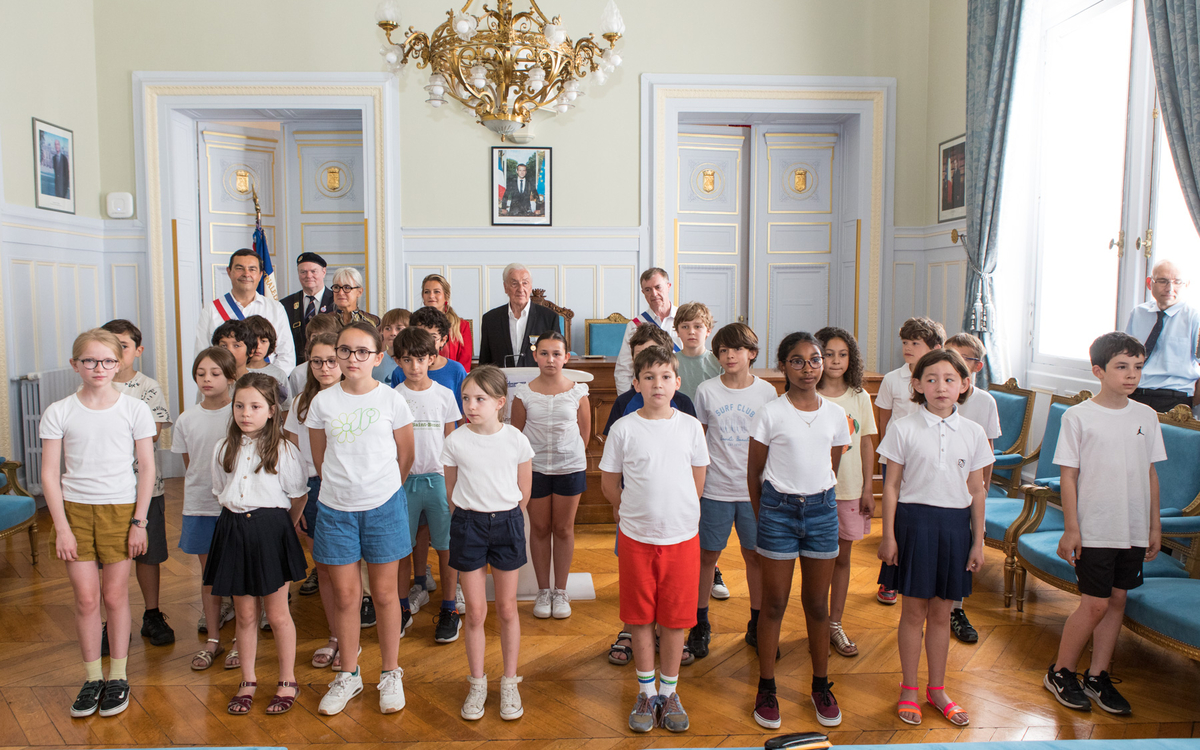 Dans la salle des fetes de la mairie, un groupe d'enfants se tient debout face à l'assemblée de la cérémonie. ils se tiennent face a la caméra. Derrrière eux plusieurs adultes dont Jean Pierre Lecoq et Moonsieur Solère assistent a l'évènement. La scène se déroule dans un salon d'apparat aux murs clairs, éclairé par de grandes fenetres et orné d'un lustre 