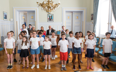 Dans la salle des fetes de la mairie, un groupe d'enfants se tient debout face à l'assemblée de la cérémonie. ils se tiennent face a la caméra. Derrrière eux plusieurs adultes dont Jean Pierre Lecoq et Moonsieur Solère assistent a l'évènement. La scène se déroule dans un salon d'apparat aux murs clairs, éclairé par de grandes fenetres et orné d'un lustre 