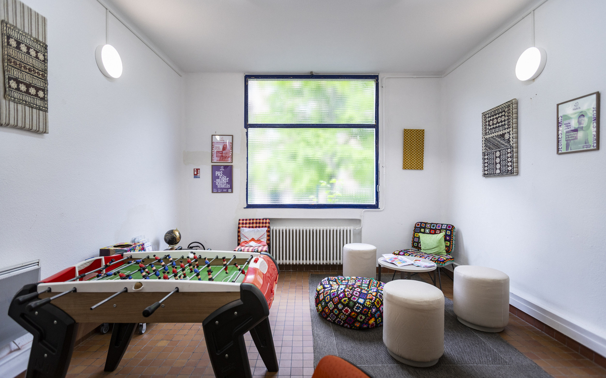 Intérieur d'une salle avec fenêtre sur végétation au fond, babyfoot au 1er plan à gauche et poufs et assises basse autour d'une table basse à droite, cadres photos aux murs