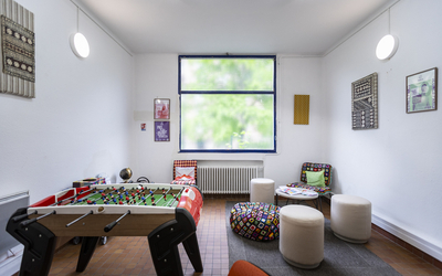 Intérieur d'une salle avec fenêtre sur végétation au fond, babyfoot au 1er plan à gauche et poufs et assises basse autour d'une table basse à droite, cadres photos aux murs