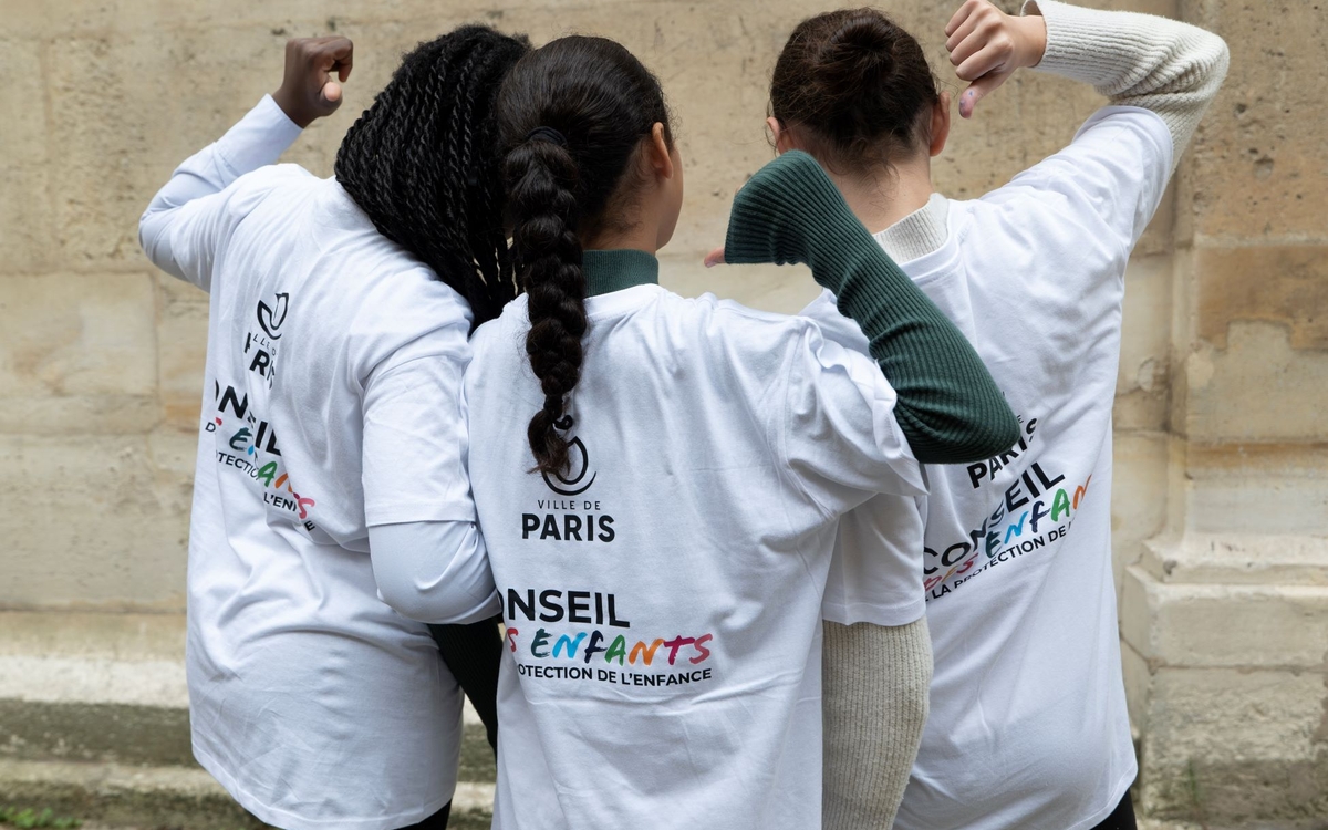 rois enfants photographiés de dos se tiennent côte à côte. Ils portent des tee-shirts avec l’inscription « Conseil des enfants » visible dans leur dos. La scène évoque la participation des jeunes à une instance citoyenne.