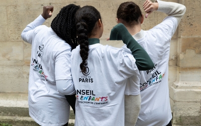 rois enfants photographiés de dos se tiennent côte à côte. Ils portent des tee-shirts avec l’inscription « Conseil des enfants » visible dans leur dos. La scène évoque la participation des jeunes à une instance citoyenne.