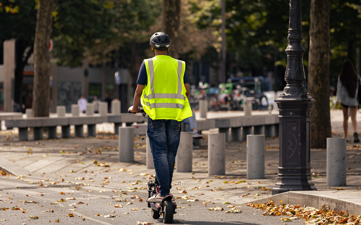 Une personne en trottinette électrique avec un casque et d'un gilet.
