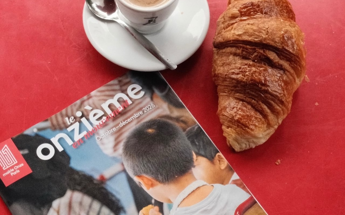 Un exemplaire du journal municipal du 11e est posé sur une table d'un café à côté d'un café et d'un croissant