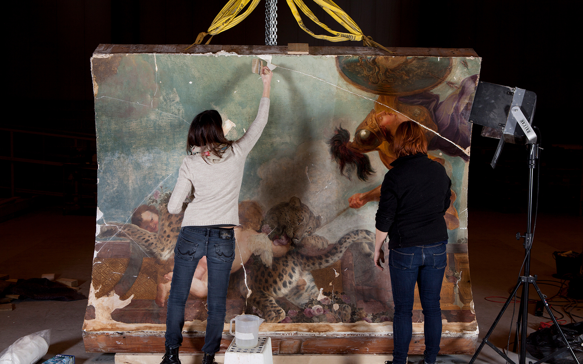 Deux restauratrices travaillent sur un fragment de plafond peint de la Chancellerie d’Orléans, suspendu par des sangles dans un atelier de restauration.