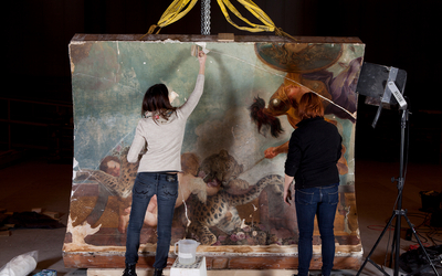 Deux restauratrices travaillent sur un fragment de plafond peint de la Chancellerie d’Orléans, suspendu par des sangles dans un atelier de restauration.
