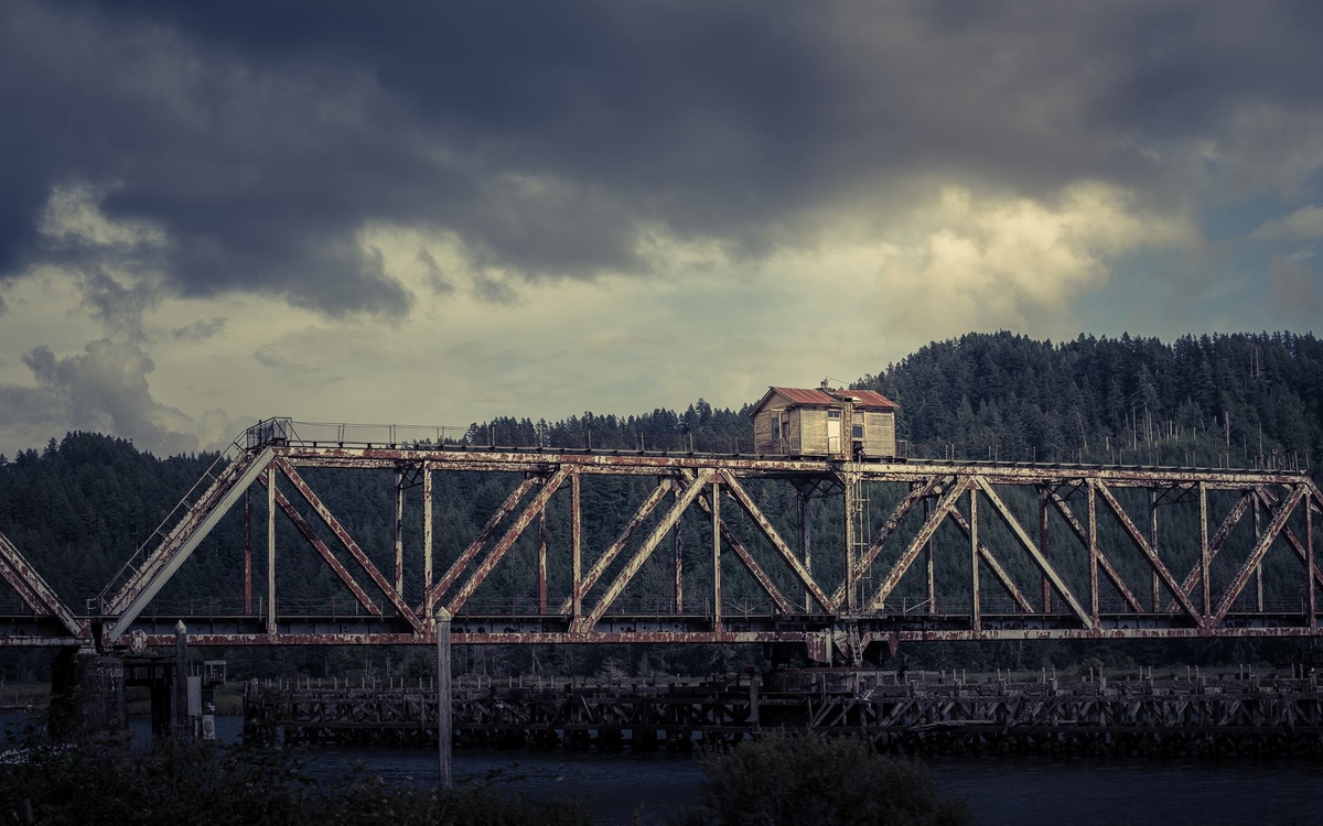 Photo d'un vieux pont industriel au dessus d'un fleuve. Une cabane est sur le pont, entre les deux rives. Au loin, on voit une forêt.
