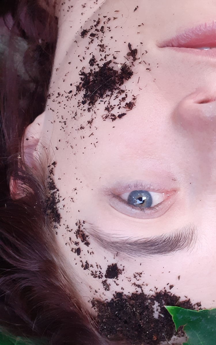 Photo du visage d'une jeune femme allongée au sol, à l'envers par rapport à la prise de vue. Son oeil gauche est visible, il est d'un bleu puissant. Sur son visage, il y a un peu de terre et deux feuilles vertes sont visibles.