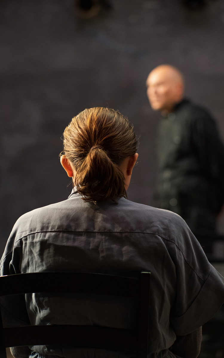 Photo d'une personne aux cheveux longs, attachés en queue de cheval. Cette personne est assise sur une chaise, de dos. En arrière plan, flou, un homme chauve, les mains dans le dos, se tient.