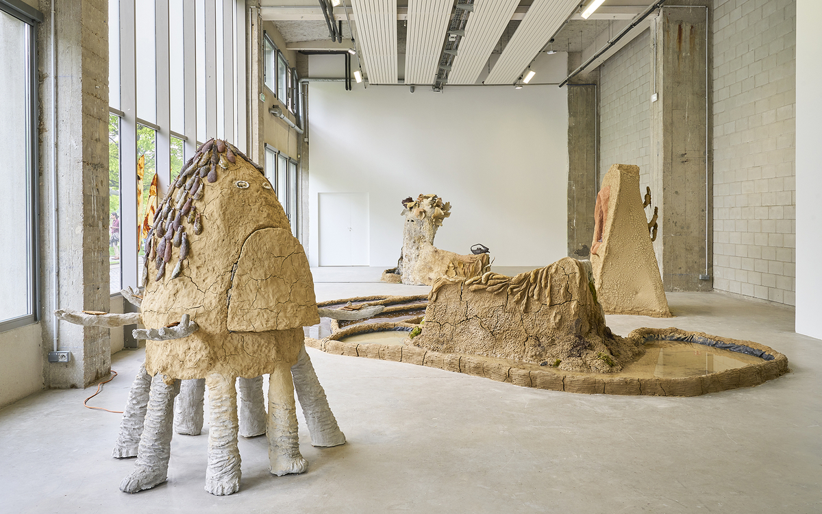 Vue d'une exposition composée de sculptures en terre crue et en béton: une fontaine, un banc, de petits étangs, etc.