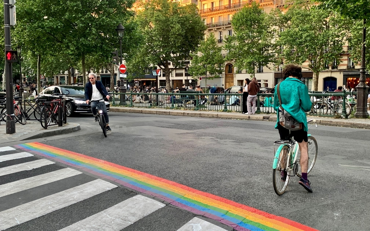 Deux personnes font du vélo dans une rue de Paris, près d'un passage piéton à côté duquel est peinte une bande aux couleurs du drapeau LGBT.
