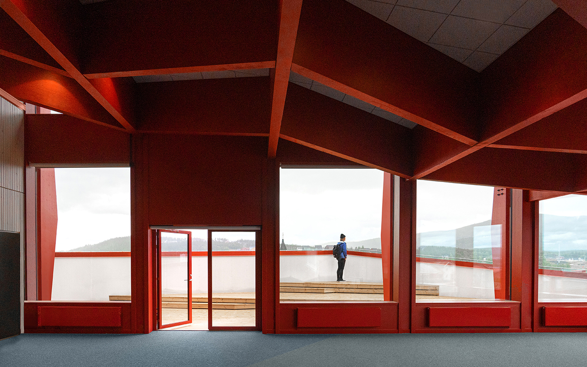 Photographie représentant l'intérieur d'un bâtiment rouge à l'architecture géométrique. En face on voit un homme à travers de grandes baies vitrées.