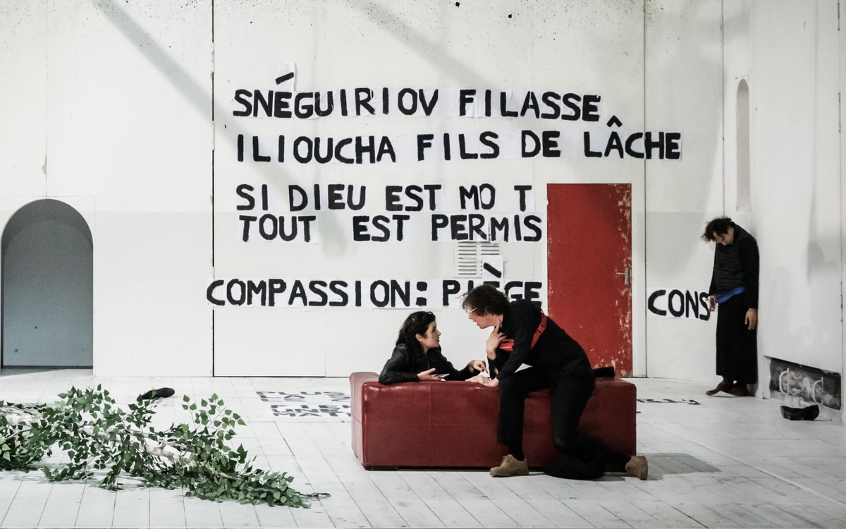 2 personnes au centre de la scène sur un canapé rouge, habillé tout de noir et discutant. Un homme, plus loin sur scène qui regarde le sol.