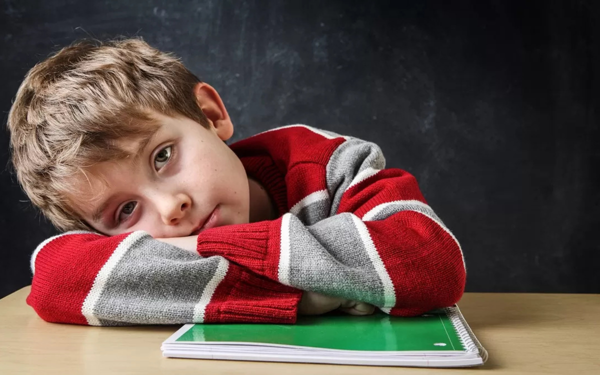 Enfant tête sur ses bras, assis à une table, nous regarde et semble s'ennuyer. 