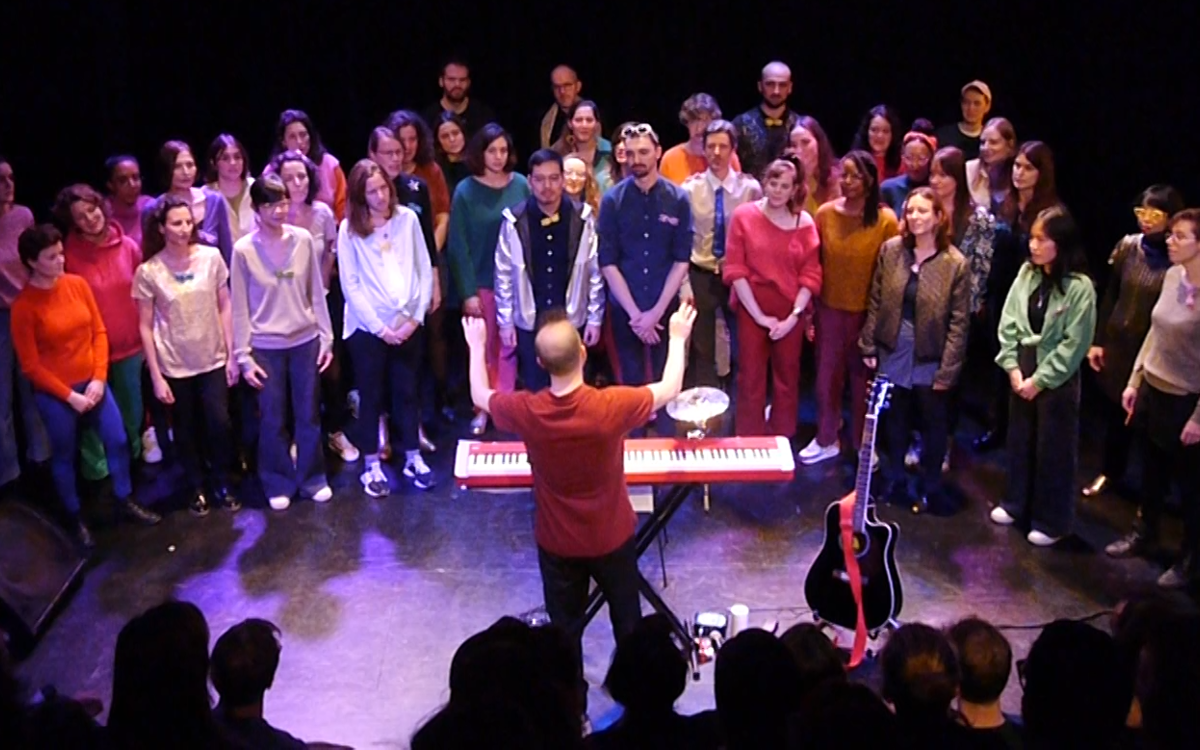 Un chef de choeur, avec son piano rouge et son cymbale est en train de diriger une chorale de 40 personnes.  Les choristes, majorité femmes, sont habillées avec plein de couleurs différents et des paillets.