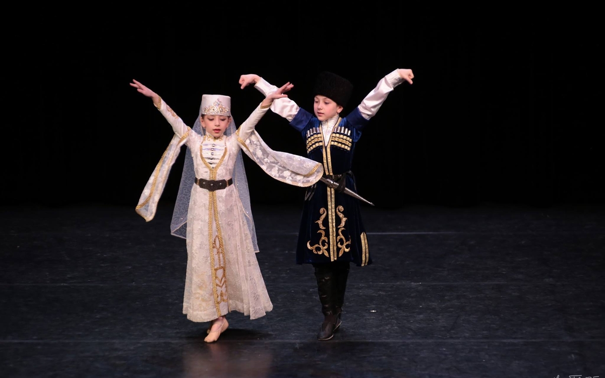 Deux enfants, en tenue traditionnelle caucasienne, dansent les bras levés seules sur une scène noir. 