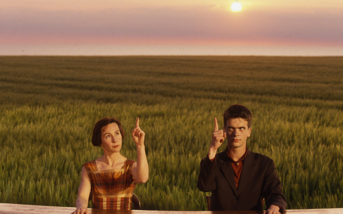 Un couple assis côte à côte lèvent leurs index vers le ciel. En arrière plan un champ.