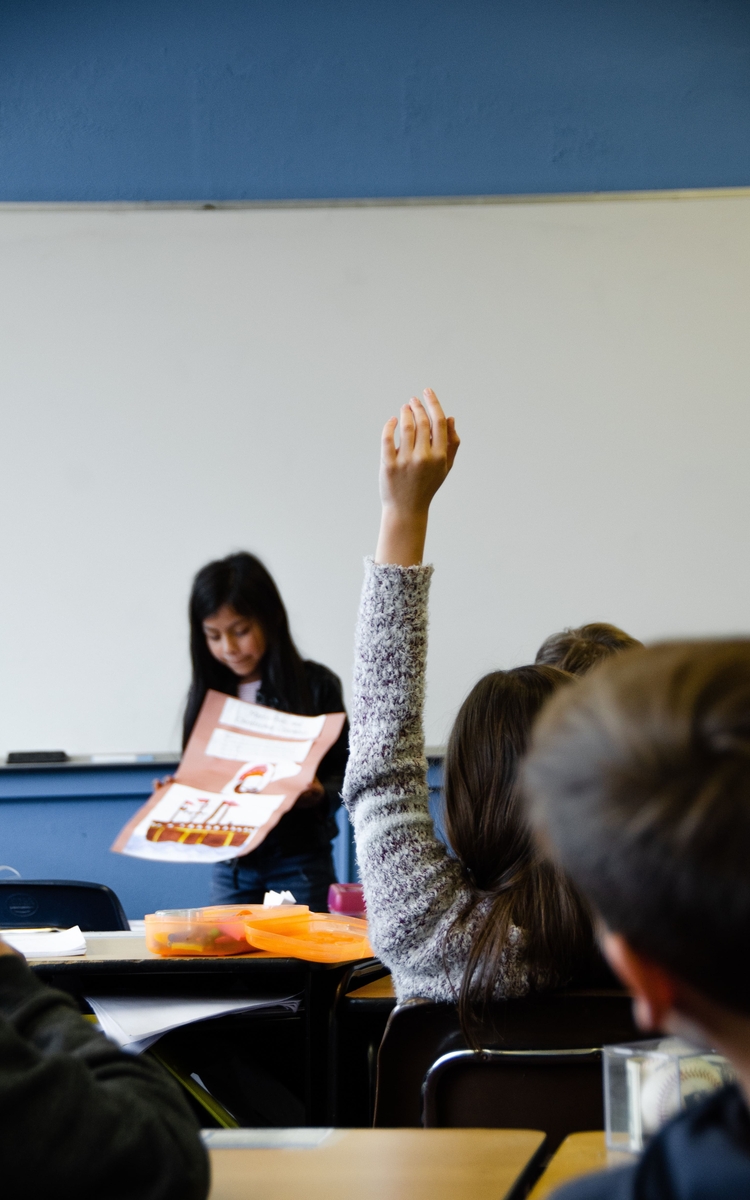 Une enfant, de dos, dans une classe d'école, lève la main. On voit en face du tableau une autre enfant exécutant un exposé.