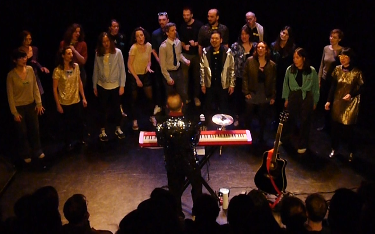 Une chorale de 20 personnes, dresscode très pailleté, entrain de chanter et danser. Le chef dirige la chorale avec son clavier rouge.