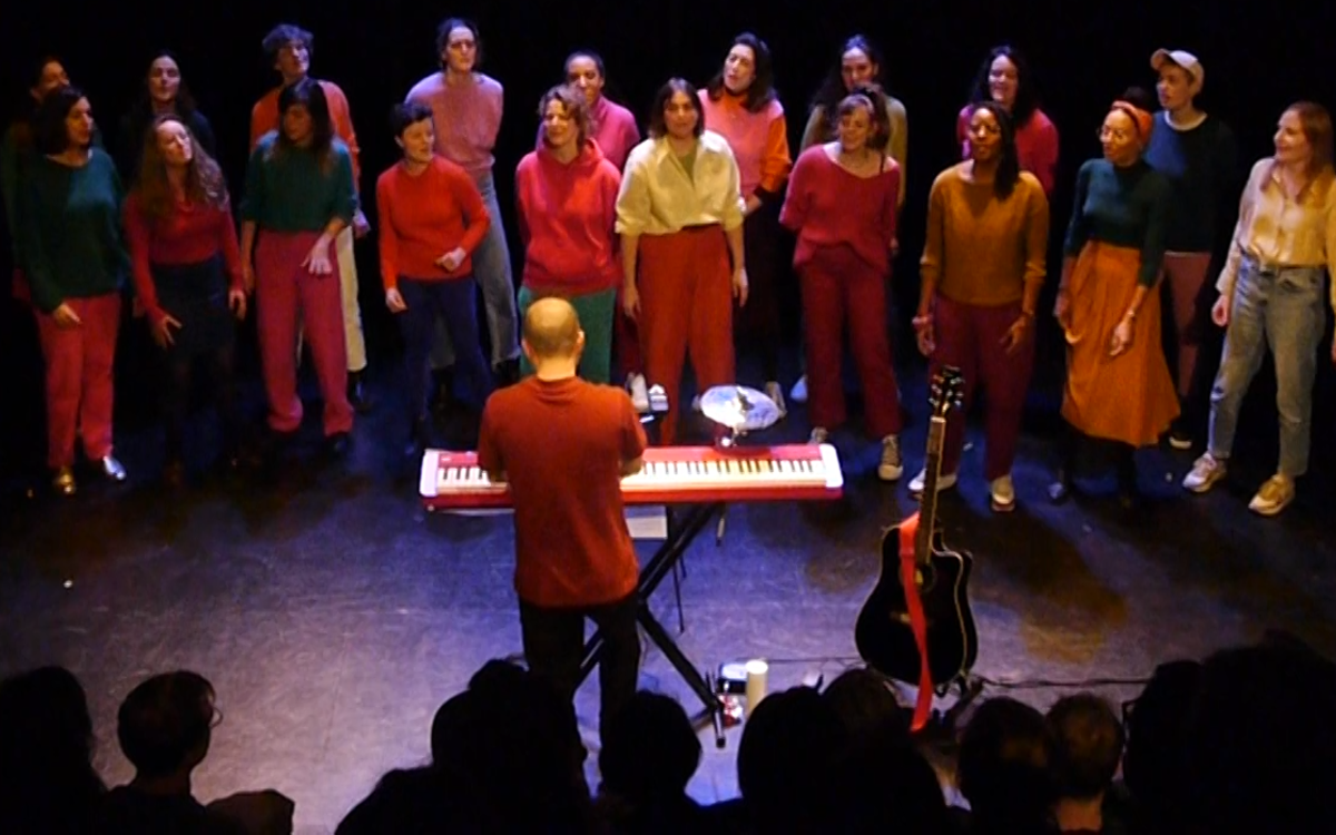 Une chorale de 20 femmes, habillées en color blocs, est entrain de chanter dans un théâtre, dirigée par un pianiste.