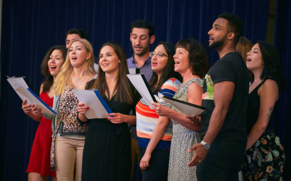 groupe de personnes, hommes et femmes de toutes origines, en train de réciter des paroles de chanson en groupe, debout sur une scène. 