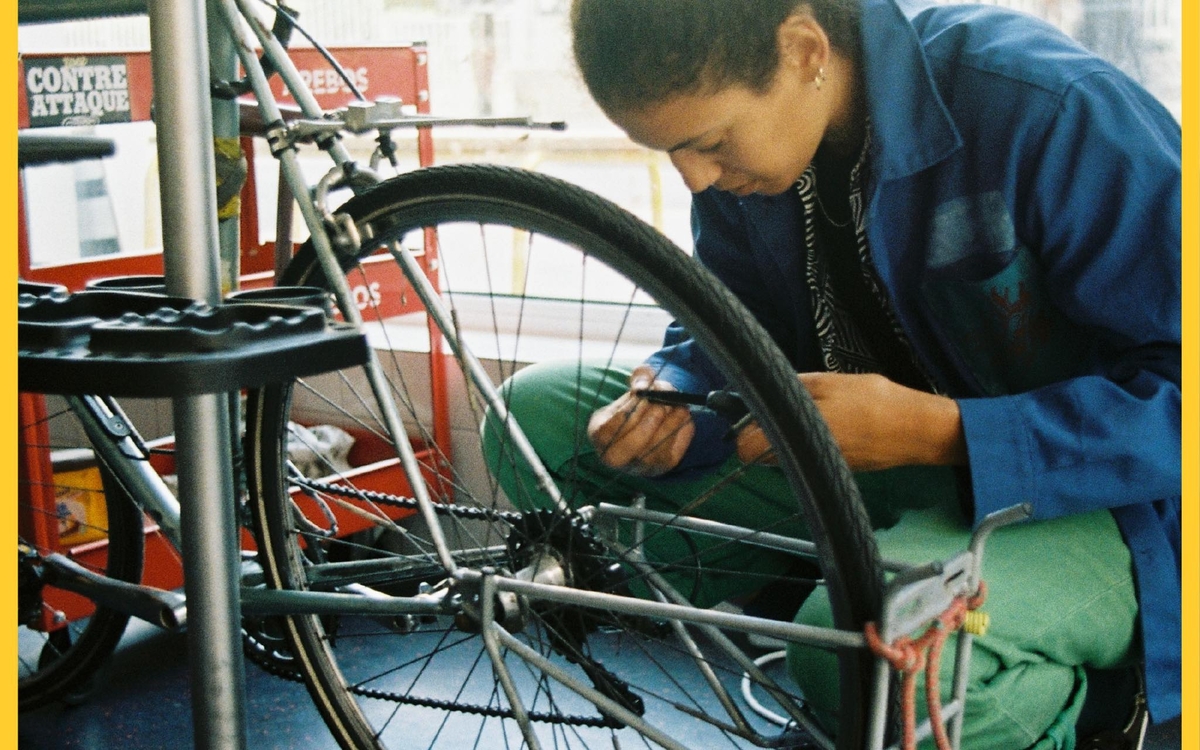 Personne métisse accroupie, les cheveux attachés, vêtue d'un bleu de travail, penchée sur un outil devant un vélo accroché sur un pied dans un ateliersur 