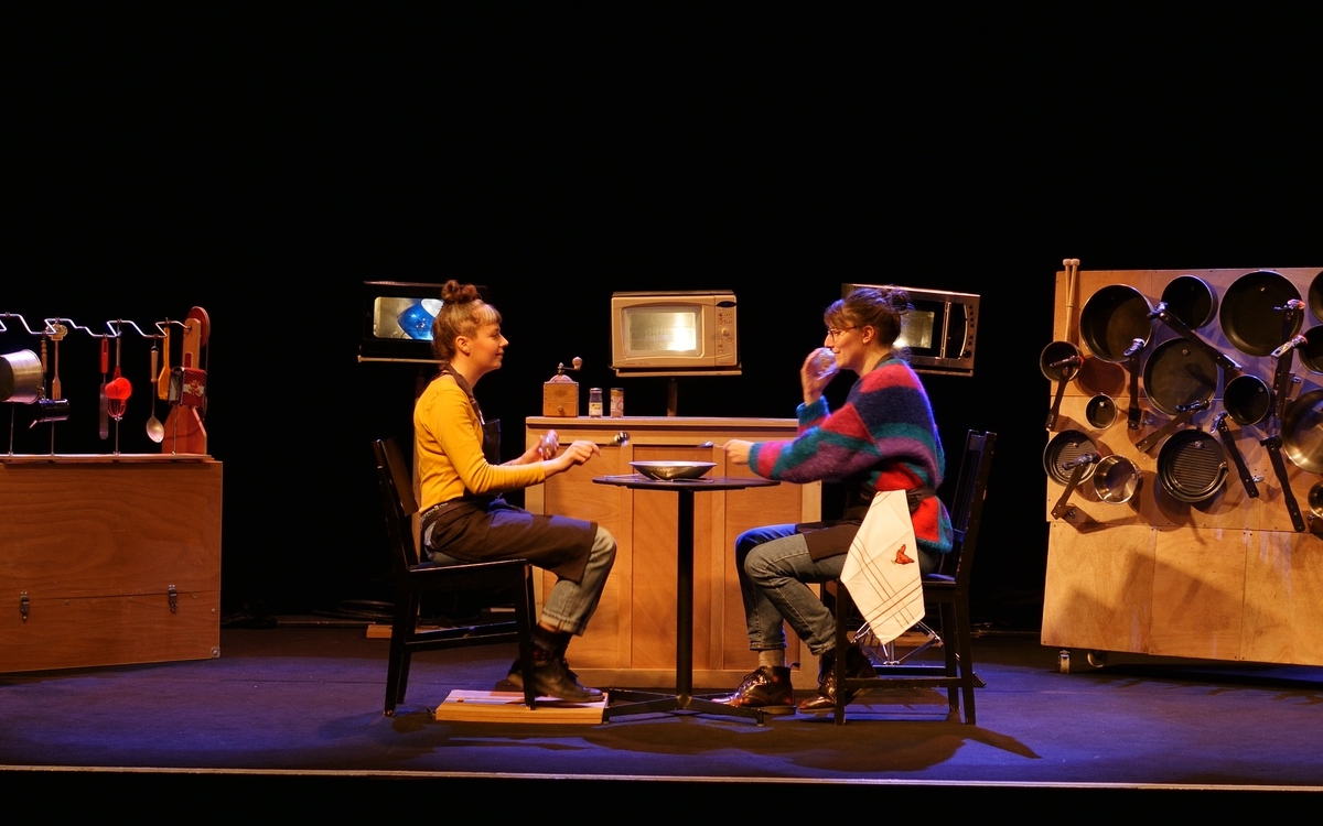 2 personnes assises à une table avec en fond de scène 3 micro-ondes, une batterie de cuisine et divers ustensiles de cuisine. 