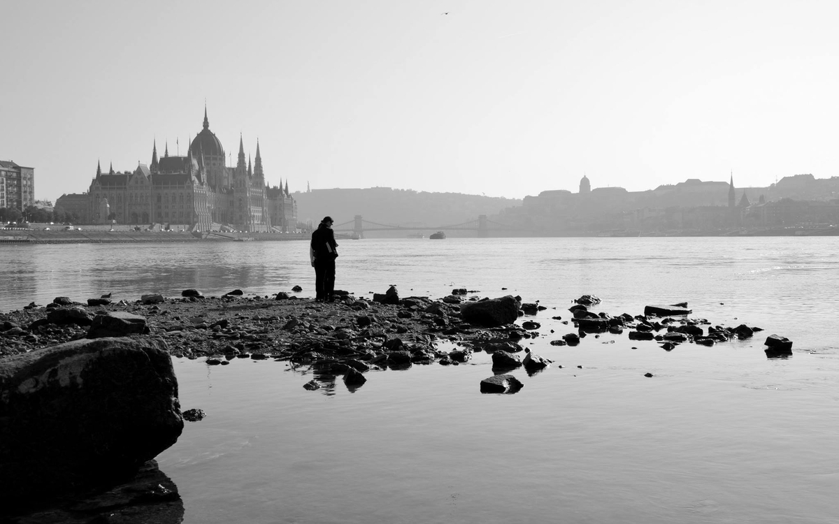 Un homme sur une grève de galet, au bord d'un lac, avec pour fond une ville de Hongrie. La photo est mélancolique, calme.
