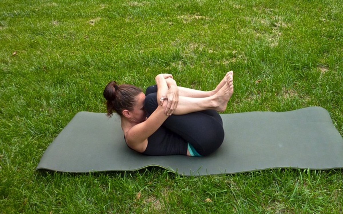 Femme allongée sur un tapis de yoga posé sur l'herbe. Posture de détente  jambes repliées vers la tête soulevée du sol.