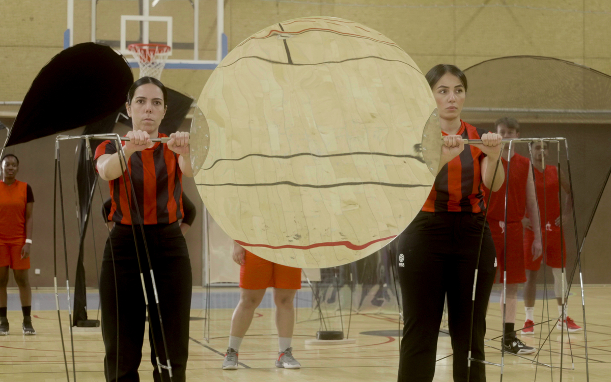 Deux arbitres tiennent frontalement une sculpture miroir de l'artiste Julia Borderie pendant un match de basket.