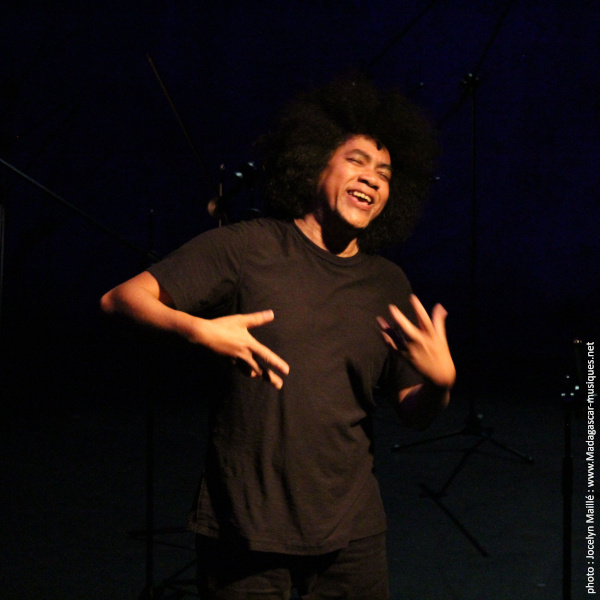 Photo du poète Jean-Luc Raharimanana souriant, les yeux fermés, levant les mains dans un halo de lumière douce, sur un plateau de spectacle 