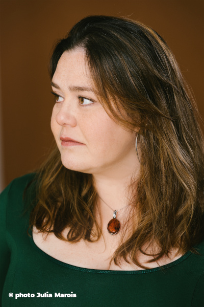 Photo de la poétesse Amélie Prévost, regardant vers sa droite. Elle a les cheveux chatains mi longs, un collier avec une pierre ambrée autour du cou et un pull vert