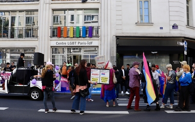 Une camionnette devant le centre LGBTQI+ de Paris IDF+ et un attroupement festif de personnes, arborant des drapeaux bi ou pan, des pancartes et une banderole aux couleurs des drapeaux bi et pan.