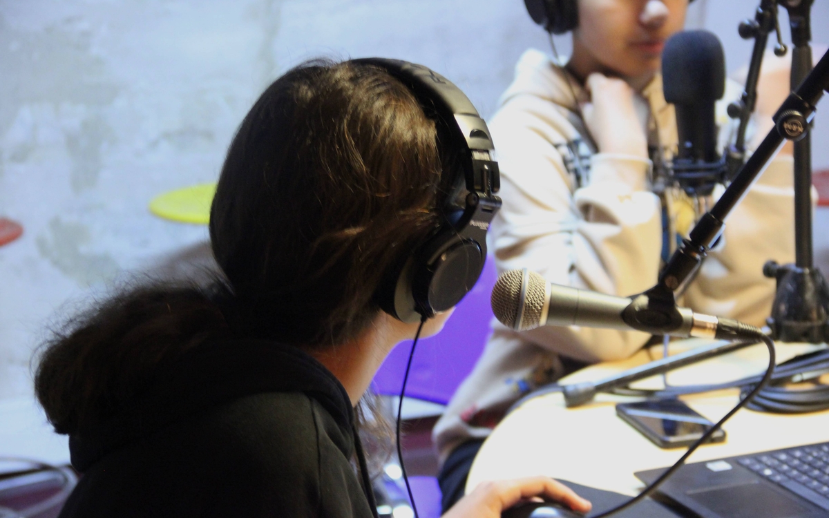 Une adolescente face à un micro radio. Dans l'arrière plan, une autre adolescente est elle aussi derrière un micro est écoute ce qui se dit.