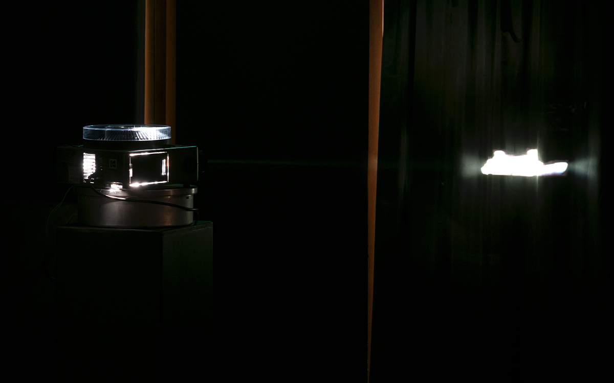 L'image est une photo de l'auditorium de Transfo plongé dans l'obscurité avec simplement une machine à diapositives qui tourne sur elle-même projetant sur le rideau d'une longue fenêtre l'image en négatif d'un bateau