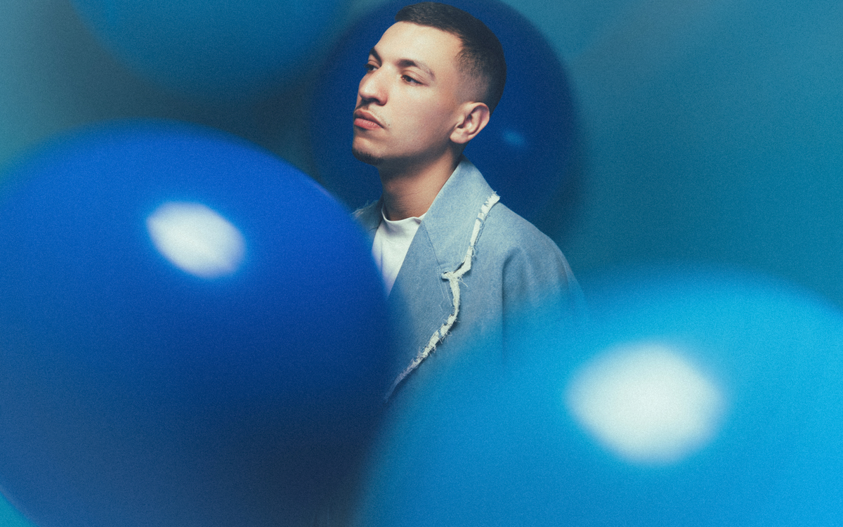 Youssef Swatt de profil devant un fond bleu et derrière des ballons bleus