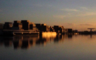 Image floue d'un paysage urbain au bord d'un fleuve au coucher de soleil, avec des reflets orangés et sombres. 