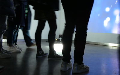 Visuel montrant un groupe dont on ne voit que les pieds devant un écran dans un espace d'exposition
