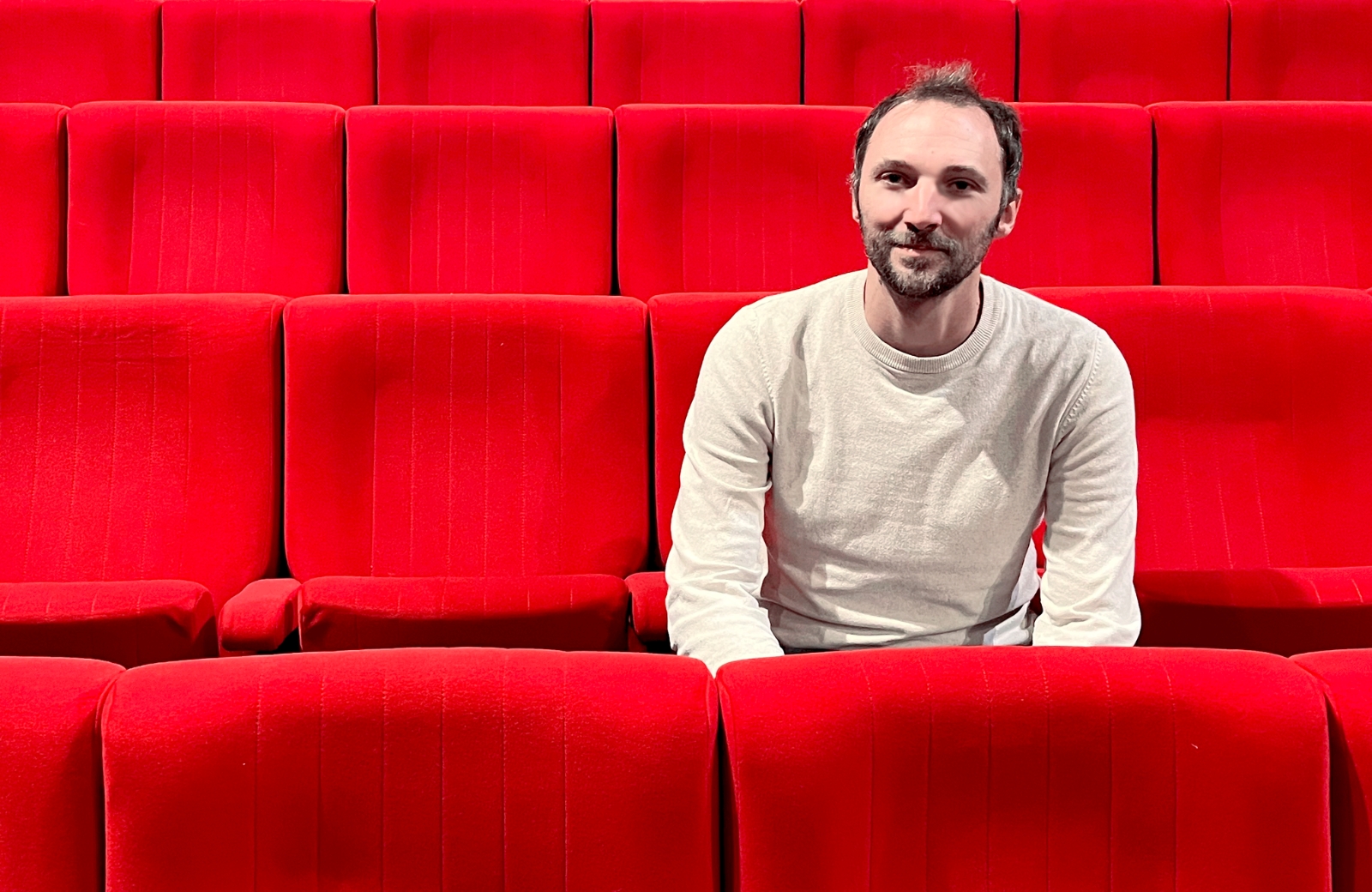 Martin Dumont assis dans un théâtre aux fauteuils rouges regarde la caméra et sourit.