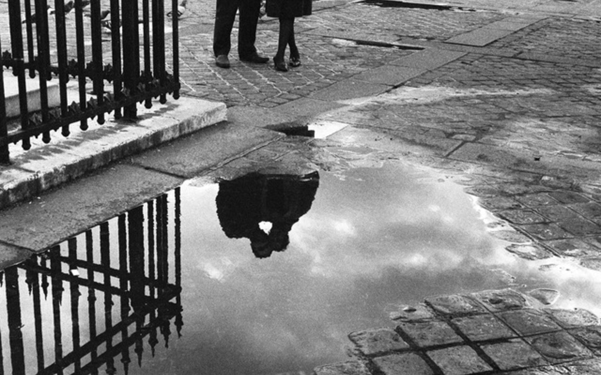 Photo en noir et blanc représentant une flaque d'eau sur des pavés où l'ombre de deux silhouettes enlacées se reflète