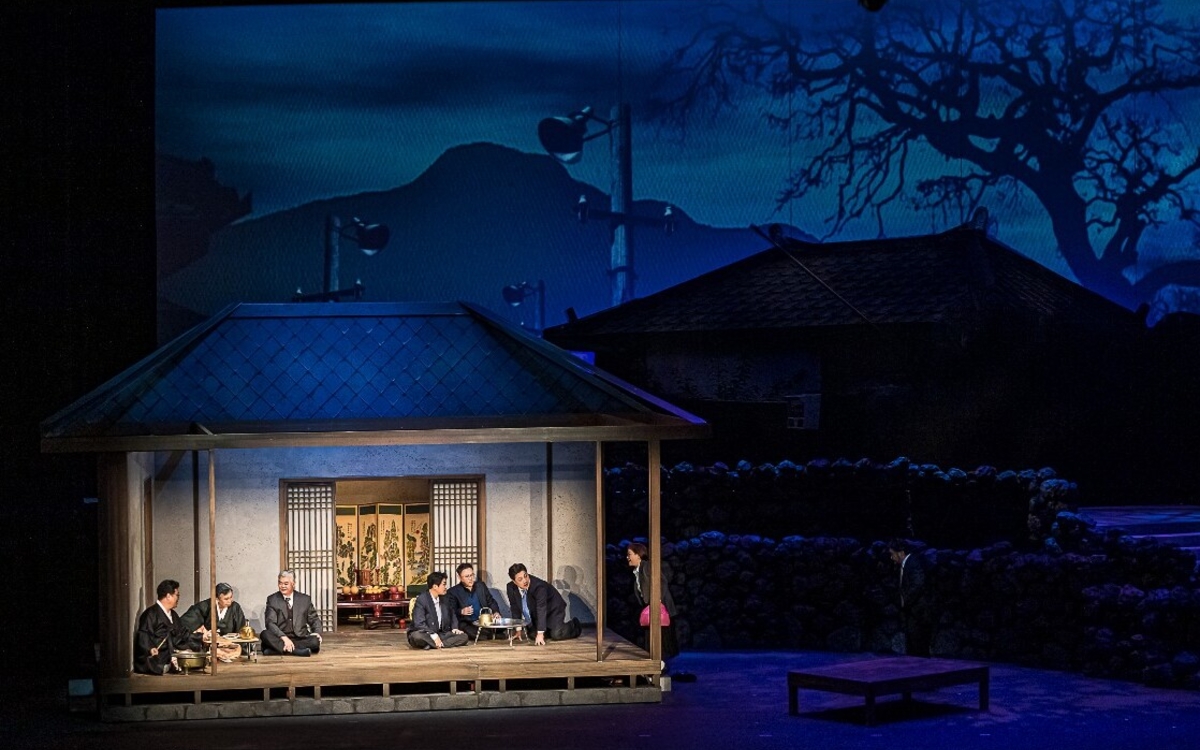 Image de l'opéra "Tante Sunni" où l'on y voit des maisons anciennes dans un paysage de nuit. Une discussion sérieuse semble s'y tenir entre une dizaine de personnages