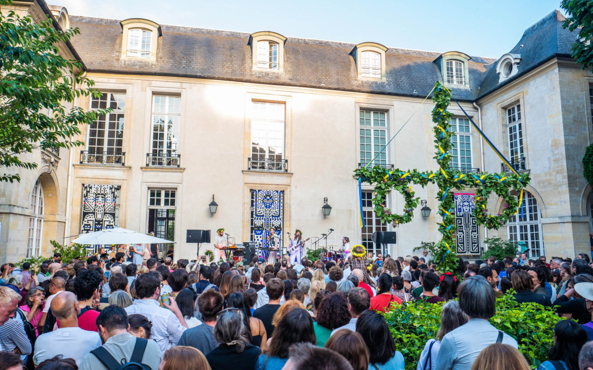 Foule rassemblée dans le jardin de l'Institut suédois, autour du mât fleuri, face à une scène où se déroule un concert.