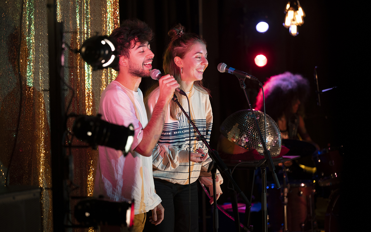 Un homme et une femme chantent, comme dans un karaoké, devant un rideau à paillette